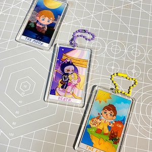 May include: Three clear acrylic keychains with colourful illustrations of tarot cards. The cards are "The Moon", "Death", and "The Fool".