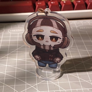 May include: A clear acrylic charm featuring a cartoon character with a skull-like face, wearing a brown hoodie, blue jeans, and headphones. The charm is attached to a small plastic stand and has a gold-colored loop at the top.