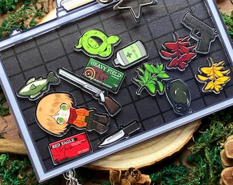 May include: A clear plastic keychain with a black and white illustration of a suitcase. The suitcase is filled with various items, including a cartoon character, a snake, a first aid kit, a gun, a knife, and a plant. The keychain also includes a small, separate charm of a cartoon character.