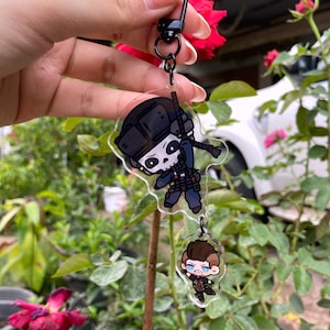 Puede incluir: Un llavero acrílico transparente con dos personajes de dibujos animados. El personaje superior es un soldado con una cara de calavera, que lleva un casco y uniforme negros. El personaje inferior tiene el pelo castaño y un atuendo marrón. El llavero tiene un cierre de metal negro.