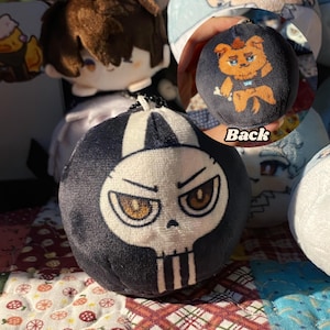 May include: A plush, round keychain featuring a skull design with a black and white color scheme. The skull has angry eyes and a stern expression. The back of the keychain shows a cartoon dog with a bone. Other plush toys are in the background.