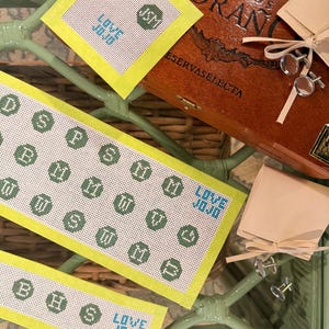 May include: Three rectangular needlepoint canvases with a light yellow border featuring green octagonal monograms, smiley faces, and the blue text LOVE JOJO. A wooden cigar box and two pairs of silver-toned circular cufflinks are also present.