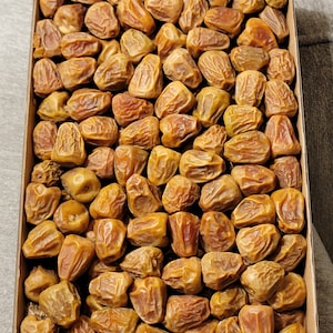 Puede incluir: Una caja rectangular de cartón llena de numerosas dátiles marrones. Los dátiles tienen una textura arrugada y un tono marrón dorado, lo que indica que están maduros. La caja se ve desde arriba, mostrando la disposición de los dátiles.