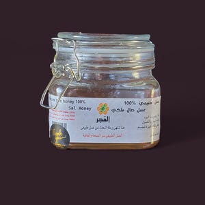 May include: A glass jar of honey with a gold lid and a label that reads "100% Natural Honey, Sal Honey, El Fajr, عسل حال ملكي 100% طبيعي".