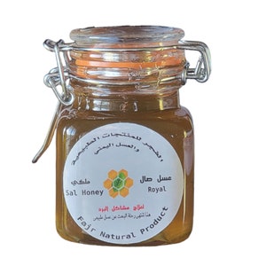 May include: A small glass jar filled with golden honey, sealed with a metal clasp and a clear lid. The jar has a white label with Arabic text and the words "Sal Honey" and "Royal". The product is labeled as a natural product.