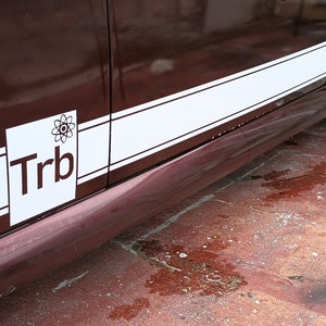 Könnte beinhalten: Ein Ausschnitt eines weinroten Autos mit einem weißen Streifen an der Seite. Ein weißes Quadrat mit den Buchstaben "Trb" und einer Grafik befindet sich auf dem Auto. Das Auto hat ein schwarzes Rad.