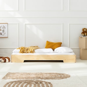 Op de afbeelding: Een houten bedframe met een wit matras, een gele deken en een geel kussen. Het bed staat in een witte kamer met een wit tapijt met bruine strepen.