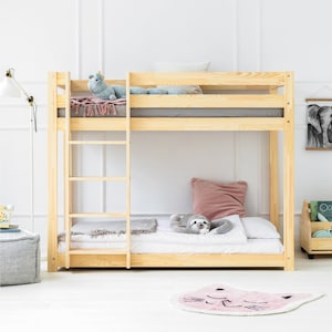 Op de afbeelding: Een houten stapelbed met een ladder aan de zijkant. Het bovenste bed heeft een lichtblauw knuffeldier en een roze deken. Het onderste bed heeft een roze kussen en een grijs knuffeldier. Er ligt een roze vloerkleed met een kattenkop op de vloer.