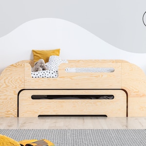 Könnte beinhalten: Ein helles Holz-Kinderbett mit einem weißen Laken mit schwarzen Punkten und einem gelben Kissen. Das Bett hat eine integrierte Schublade und ein Sicherheitsgitter.