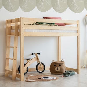 Peut inclure: Un lit superposé en bois avec une échelle et une barre de sécurité intégrée. Le lit se trouve dans une chambre d'enfant avec un tapis vert et marron, un vélo en bois, un animal en peluche et un panier de rangement avec un visage d'ours.