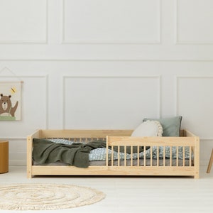 Puede incluir: Una cama de madera clara para niños pequeños con una manta verde y una sábana con estampado azul y blanco. La cama tiene una barandilla de seguridad en un lado. Hay una alfombra en el suelo, una pequeña mesa de madera con un teléfono de juguete encima y una lámpara de pie con una pantalla blanca.