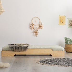 Puede incluir: Una cama plataforma de madera con un colchón verde claro y una manta de lana gris. La cama está en una habitación con una pared blanca, un atrapasueños y una alfombra en blanco y negro.