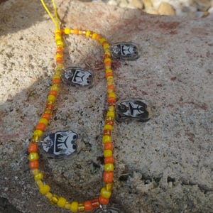 Könnte beinhalten: Ein mit gelben, orangefarbenen und transparenten Perlen besetztes Schlüsselband. Das Schlüsselband hat mehrere kleine, transparente Anhänger mit einem schwarz-weißen Design. Es liegt auf einer rauen, grauen Steinoberfläche.