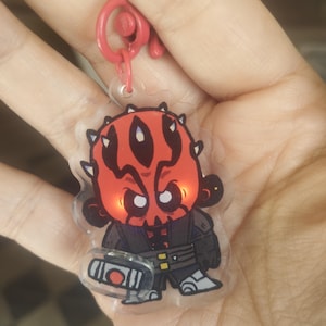 Puede incluir: Un llavero acrílico transparente con una representación de dibujos animados de Darth Maul. El personaje tiene una cara roja con marcas negras, vistiendo un atuendo negro. El llavero tiene un cierre de plástico rojo.