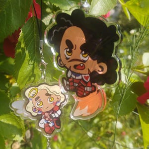 May include: Two cartoon character acrylic charms hanging from a black cord and a silver chain. One charm features a character with black hair and a red and white outfit. The other charm has a character with blonde hair and a red and white outfit.