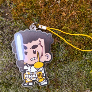 Pode incluir: Um chaveiro de acrílico transparente com um personagem de desenho animado segurando um sabre de luz. O personagem tem cabelo castanho, um traje branco e amarelo e uma cicatriz no rosto. O chaveiro tem uma corda amarela.