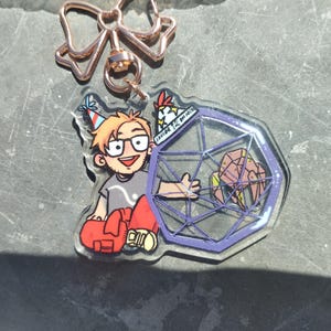 May include: An acrylic keyring featuring a cartoon character with blonde hair, glasses, and a party hat, sitting next to a purple geometric shape containing a small brown creature. The keyring has a rose gold bow-shaped clasp at the top.