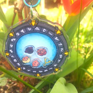 May include: A clear, circular keychain with a Stargate design. It has a blue centre with cartoon faces and a black and yellow border with symbols. A silver-coloured, iridescent key ring is attached.