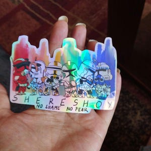 May include: A holographic sticker featuring a group of cartoon characters in a line. The characters are wearing helmets and armor. The sticker has the text "SHERESHOY NO SHAME NO FEAR" at the bottom.