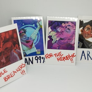 Può includere: Quattro stampe colorate in stile Polaroid con diverse illustrazioni e testi. La prima stampa presenta due persone con accenti rossi e blu, con il testo "RULE BREAKERS!!!". La seconda stampa presenta una persona con accenti blu e rosa, con il testo "AN 99%". La terza stampa presenta due persone con accenti rosa e viola, con il testo "FOR THE REPUBLIC!!". La quarta stampa presenta due persone con accenti blu e verdi, con il testo "ARCS!".