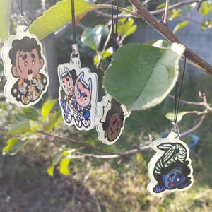 Pode incluir: Cinco amuletos de personagens coloridos pendurados em cordas pretas em um galho de árvore. Os amuletos são feitos de acrílico transparente e apresentam diferentes personagens de desenhos animados.