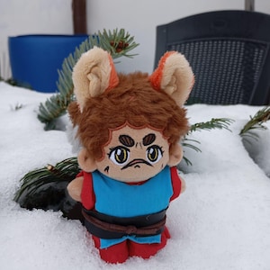 Pode incluir: Um brinquedo de pelúcia com pelo marrom, orelhas laranja e uma roupa azul e vermelha. O brinquedo tem um rosto de desenho animado com sobrancelhas marrons, um bigode e olhos grandes. O brinquedo está sentado na neve.