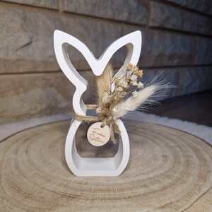 May include: A white wooden Easter bunny ornament with a cut-out design. It's decorated with dried flowers and tied with twine. A small wooden tag with text is attached. The ornament is displayed on a wooden slice, a rustic Easter decoration.