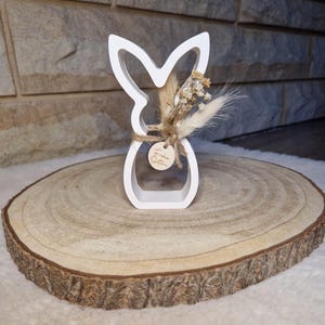 May include: A white wooden Easter bunny ornament with a floral arrangement and a small wooden tag that says "Frohe Ostern". The bunny is displayed on a round wooden slice.