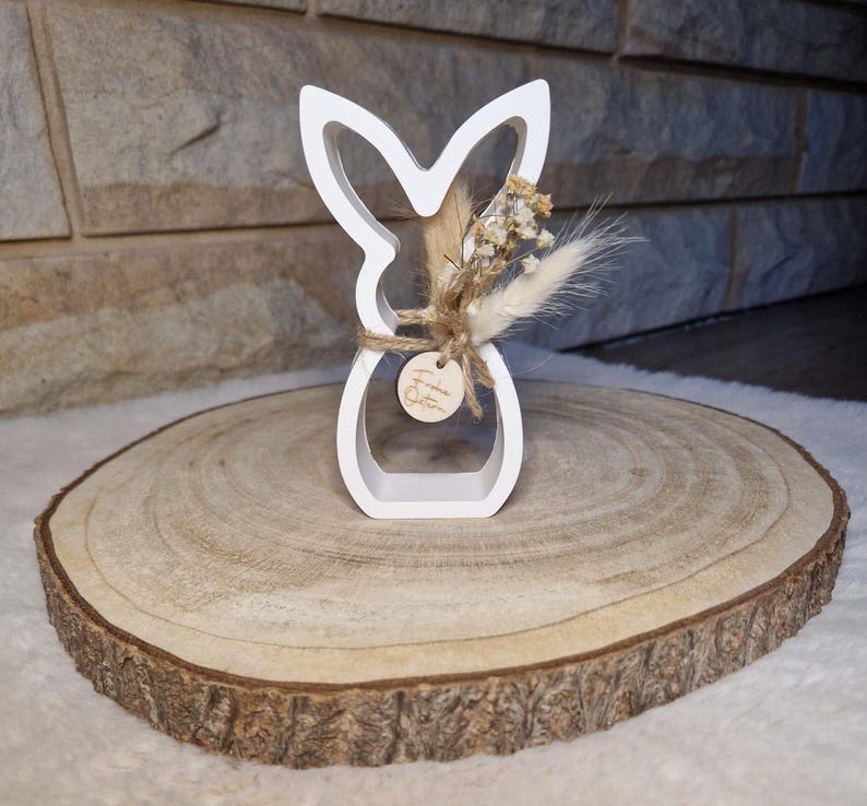 May include: A white wooden Easter bunny ornament with a floral arrangement and a small wooden tag that says "Frohe Ostern". The bunny is displayed on a round wooden slice.