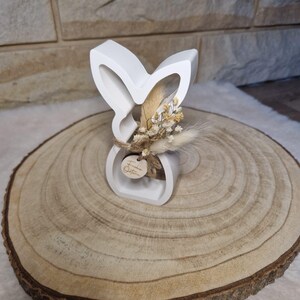 May include: A white, bunny-shaped ornament with a small bunch of dried flowers and a wooden tag. The bunny sits on a round wooden slice, showing growth rings. The tag has text on it.