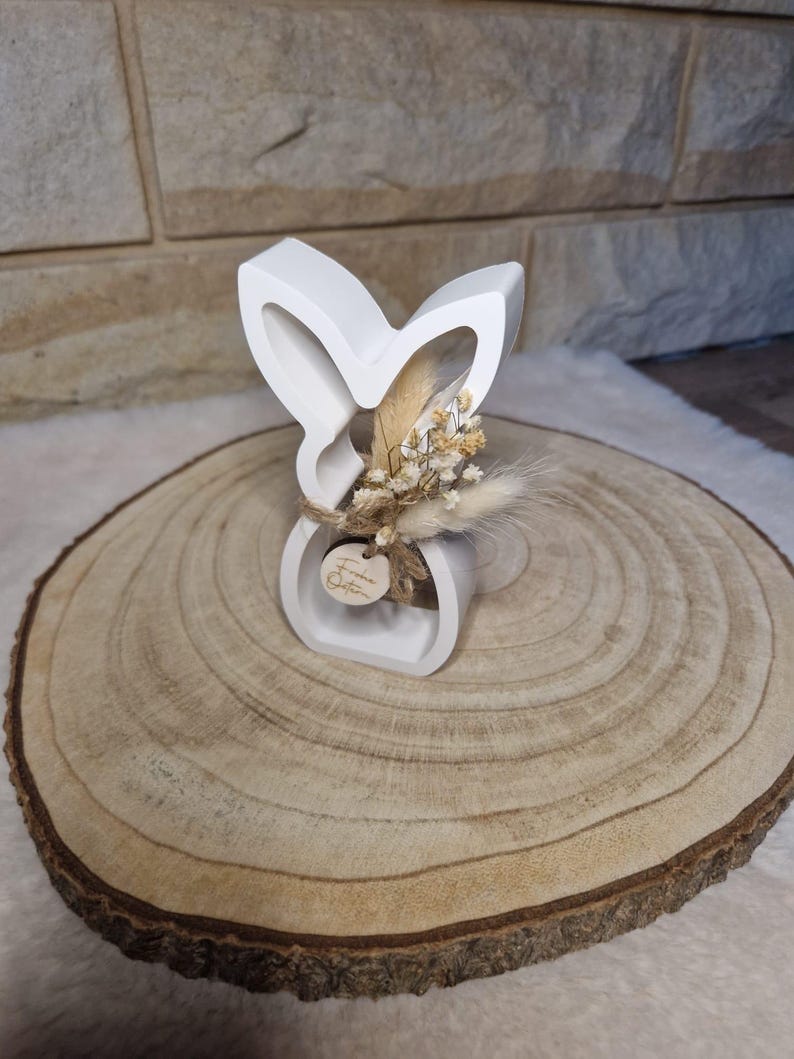 May include: A white, bunny-shaped ornament with a small bunch of dried flowers and a wooden tag. The bunny sits on a round wooden slice, showing growth rings. The tag has text on it.