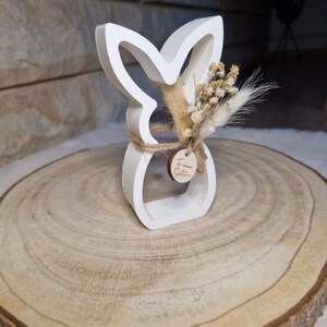 May include: A white bunny-shaped ornament with a floral arrangement and a small wooden tag. The bunny has a cutout design and is made of a solid material. The floral arrangement includes dried flowers and grasses. The ornament is tied with twine and sits on a wooden slice.