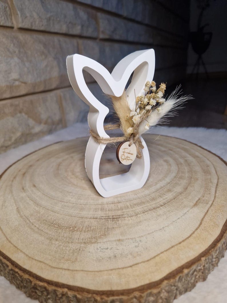 May include: A white bunny-shaped ornament with a floral arrangement and a small wooden tag. The bunny has a cutout design and is made of a solid material. The floral arrangement includes dried flowers and grasses. The ornament is tied with twine and sits on a wooden slice.
