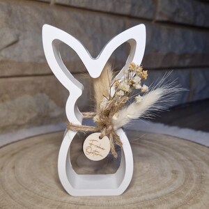 May include: A white wooden Easter bunny silhouette with dried flowers and a small wooden tag that says "Frohe Ostern". The bunny is decorated with natural-coloured dried flowers and tied with twine. The bunny is on a wooden surface.