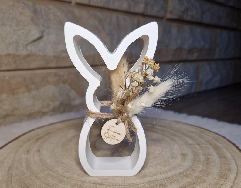 May include: A white wooden Easter bunny silhouette with dried flowers and a small wooden tag that says "Frohe Ostern". The bunny is decorated with natural-coloured dried flowers and tied with twine. The bunny is on a wooden surface.