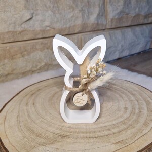 May include: A white wooden bunny-shaped ornament with a hollow centre, decorated with dried flowers and tied with twine. The bunny sits on a round wooden slice, ideal for Easter or spring decor.