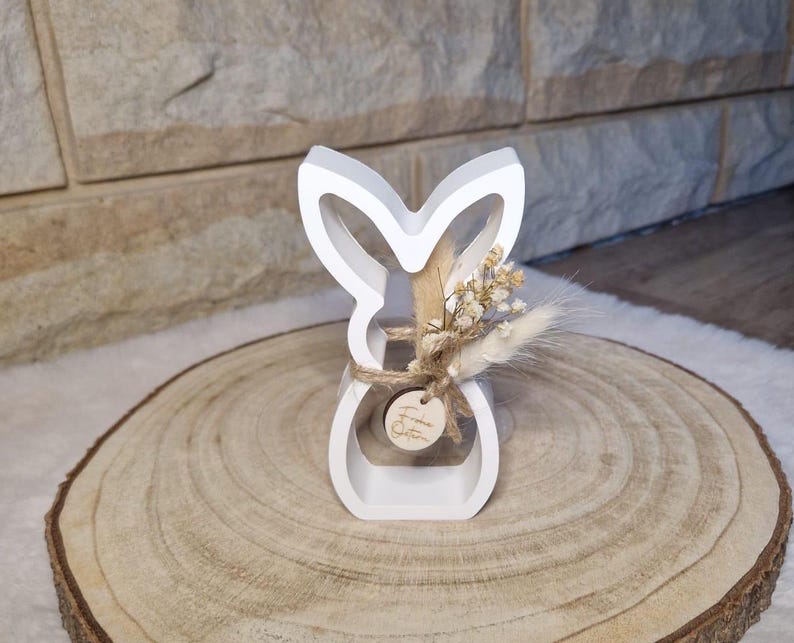 May include: A white wooden bunny-shaped ornament with a hollow centre, decorated with dried flowers and tied with twine. The bunny sits on a round wooden slice, ideal for Easter or spring decor.