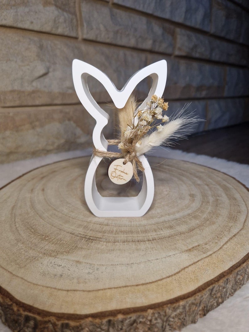 May include: A white wooden Easter bunny ornament with a cut-out design. It's decorated with dried flowers and tied with twine. A small wooden tag with text is attached. The ornament is displayed on a wooden slice, a rustic Easter decoration.