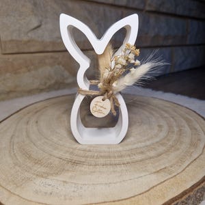 May include: A white wooden Easter bunny ornament with a cut-out design. The bunny is decorated with dried flowers and a small wooden tag tied with twine. The tag has the words "Frohe Ostern" engraved on it. The ornament is on a wooden slice.