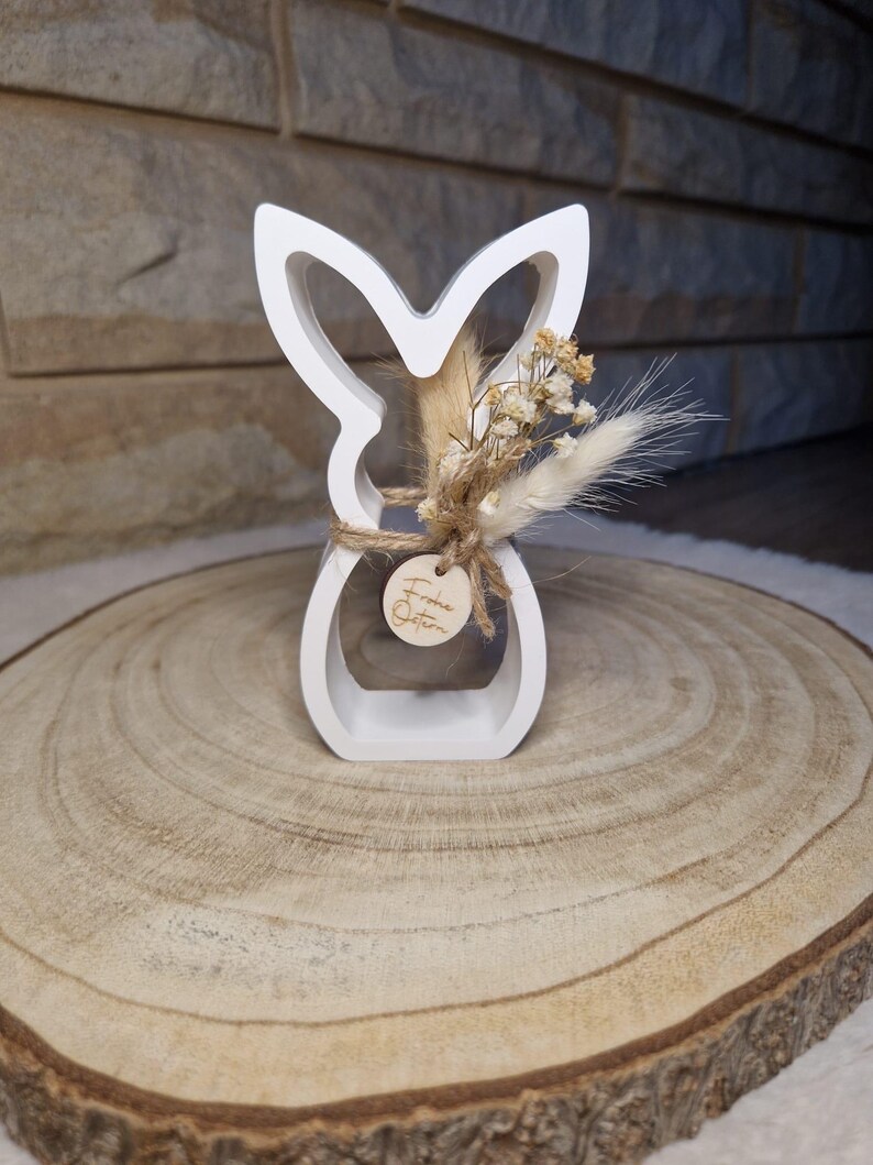 May include: A white wooden Easter bunny ornament with a cut-out design. The bunny is decorated with dried flowers and a small wooden tag tied with twine. The tag has the words "Frohe Ostern" engraved on it. The ornament is on a wooden slice.