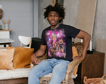May include: A person wearing a black t-shirt with a pink and white graphic print of a group of people. The shirt has the word "JOI" printed in pink letters. The person is sitting on a wicker chair with their feet on a woven rug.
