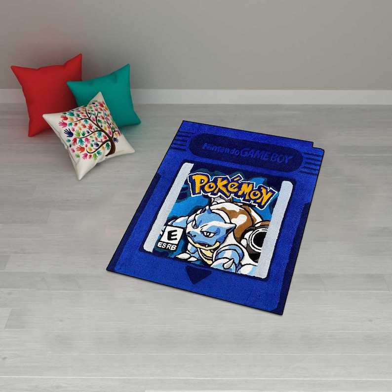 May include: A blue rectangular rug featuring the Pok&eacute;mon logo and a graphic of Blastoise, designed to resemble a Nintendo Game Boy cartridge. The rug is placed on a light-colored wooden floor, with three decorative pillows in the background.