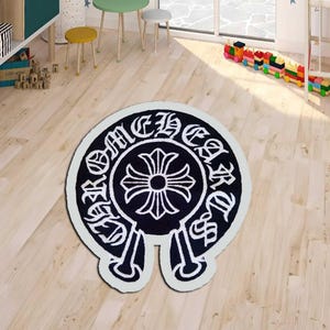 May include: A black and white area rug with a distinctive emblem design. The rug features a central cross motif surrounded by stylized text, set against a black background with a white border. The rug is placed on a light wood floor.