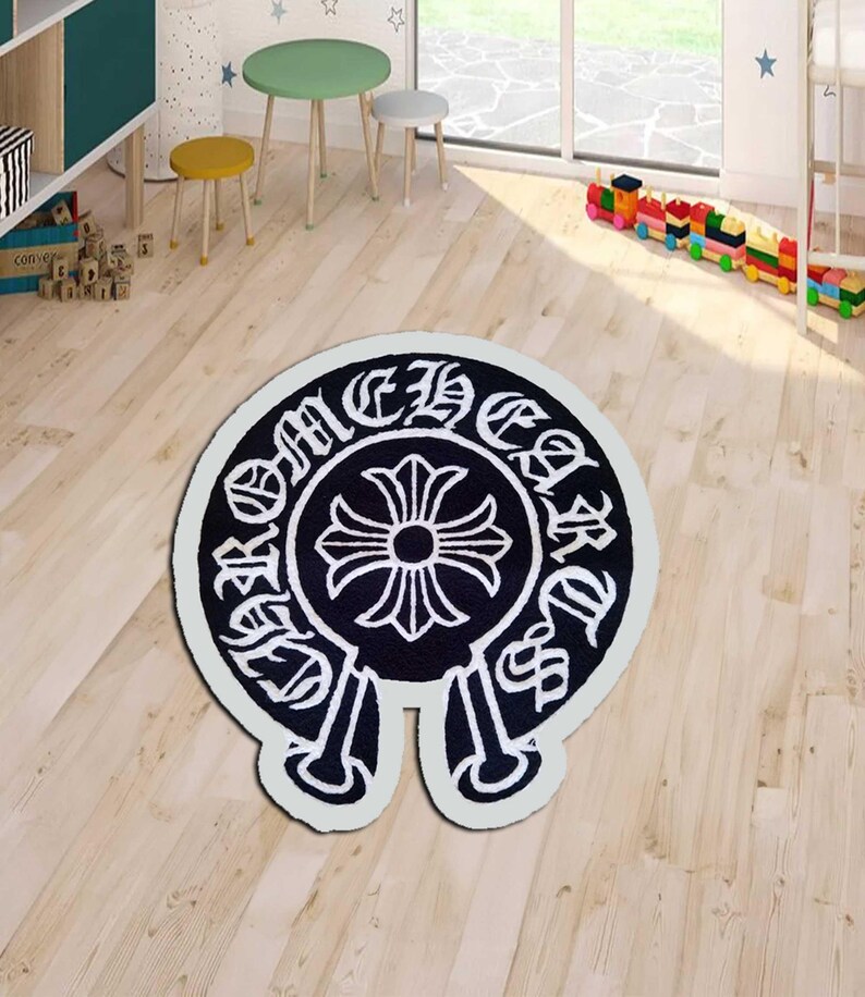 May include: A black and white area rug with a distinctive emblem design. The rug features a central cross motif surrounded by stylized text, set against a black background with a white border. The rug is placed on a light wood floor.