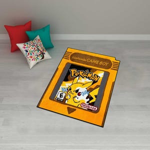 May include: A rectangular rug designed to resemble a Nintendo Game Boy cartridge. The rug features the Pok&eacute;mon logo and a yellow Pikachu character. The rug is yellow with black and gray accents and the text "Nintendo GAME BOY".