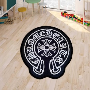 May include: Black and white round rug with a decorative emblem. The emblem features a cross design in the center, surrounded by text. The rug is on a light wood floor.