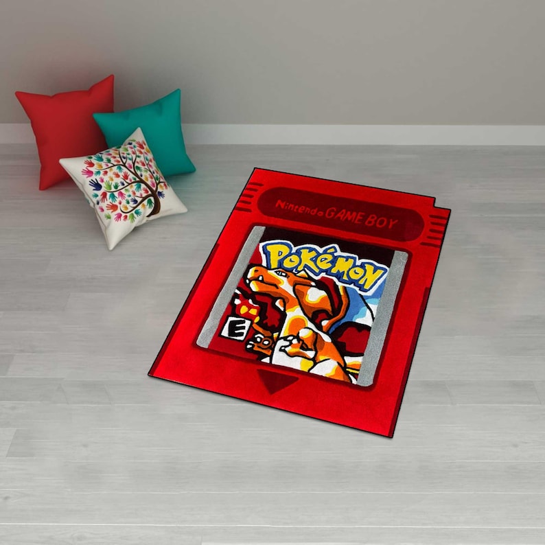 May include: A red rug shaped like a Nintendo Game Boy cartridge, featuring the Pokémon logo and a Charizard illustration. The rug is on a light gray floor, with red, teal, and patterned pillows in the background.