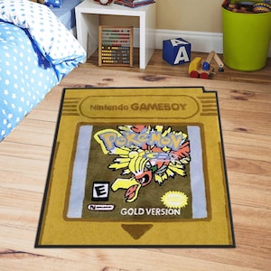 May include: A rectangular rug designed to resemble a Nintendo Game Boy cartridge. The rug features the "Pokemon Gold Version" design with a yellow and black border. The rug is placed on a wooden floor.