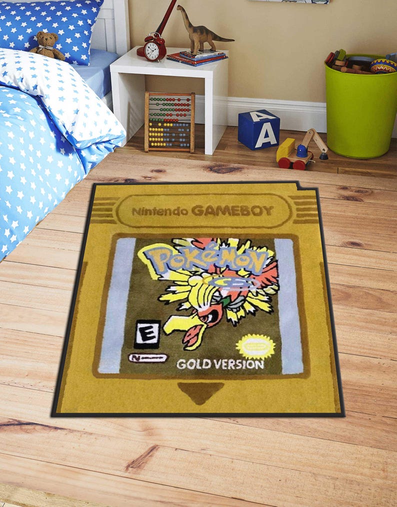 May include: A rectangular rug designed to resemble a Nintendo Game Boy cartridge. The rug features the "Pokemon Gold Version" design with a yellow and black border. The rug is placed on a wooden floor.