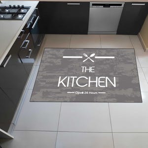 Può includere: Un tappeto rettangolare da cucina con uno sfondo grigio effetto legno invecchiato. Il testo bianco recita "THE KITCHEN" con una grafica di forchetta e cucchiaio sopra e "Open 24 Hours" sotto.